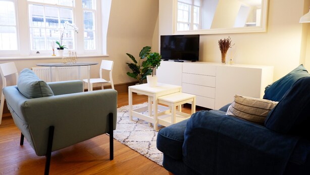 Uma sala de estar moderna com um sofá, uma mesa de centro e uma TV em um armário branco. Há uma planta na mesa de centro e uma janela com vista para o exterior.
