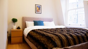Quarto com cama, mesa de cabeceira com planta, janela com vista para edifícios e quadro na parede.