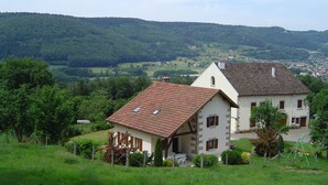 A two-story house with a sloped roof, surrounded by lush greenery and hills in the background. - Forest vacations 3 nights minimum (Le Val-d'Ajol)