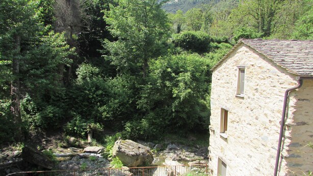 Un edificio de piedra con una ventana, rodeado de abundante vegetación y un arroyo rocoso.