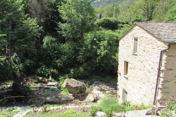 Un edificio de piedra con una ventana, rodeado de abundante vegetación y un arroyo rocoso.