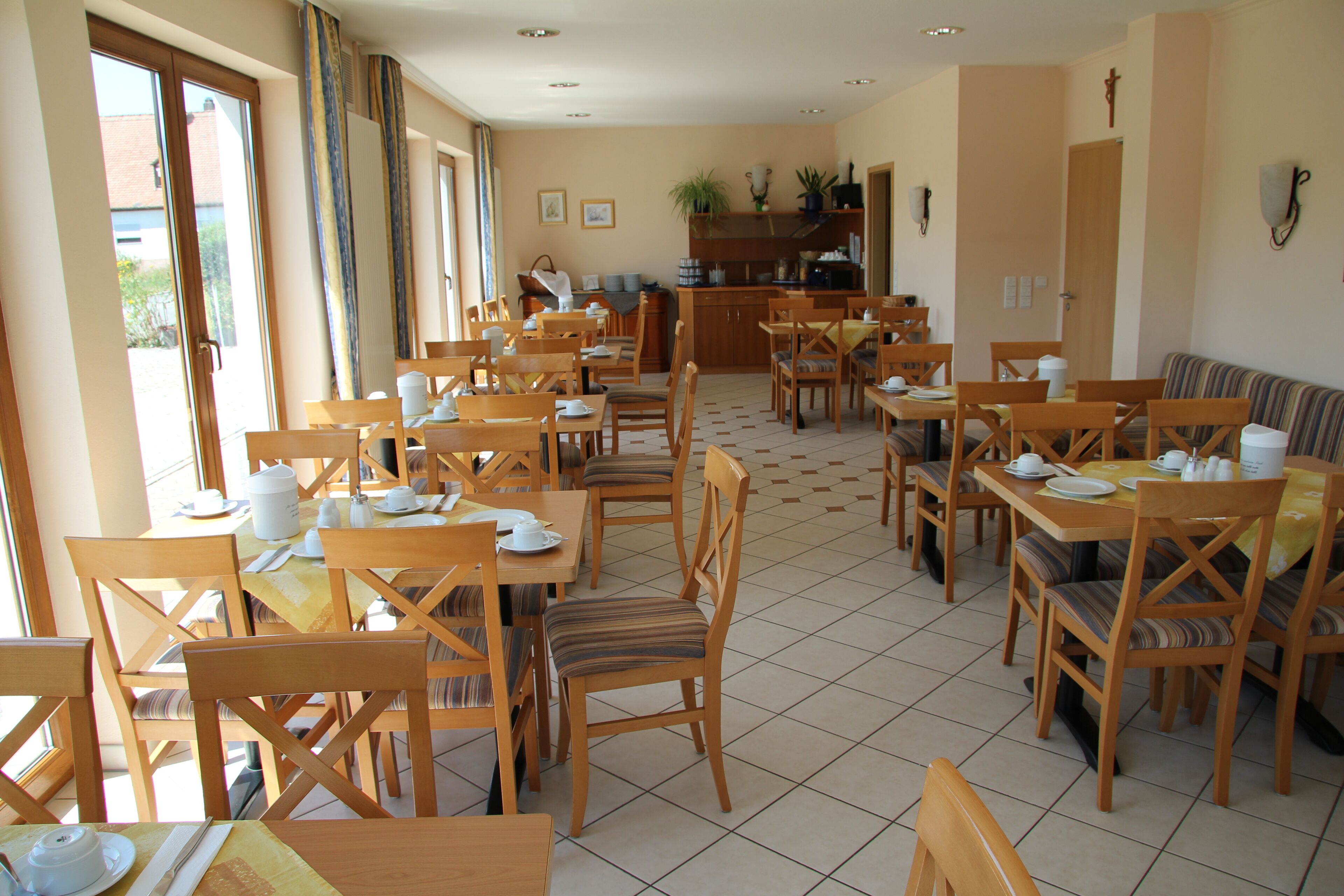 A dining area with multiple wooden tables and chairs, a kitchen in the background, and a cross on the wall.