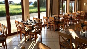 A dining area with wooden tables and chairs, large windows with a view of a green field, and a tiled floor. - Annas Gästehaus (Hallerndorf)