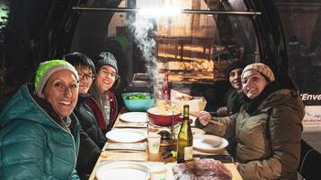 Un groupe de personnes est assis autour d’une table en bois à l’intérieur d’un véhicule doté d’un toit transparent, et profite d’un repas composé de différents plats et boissons.