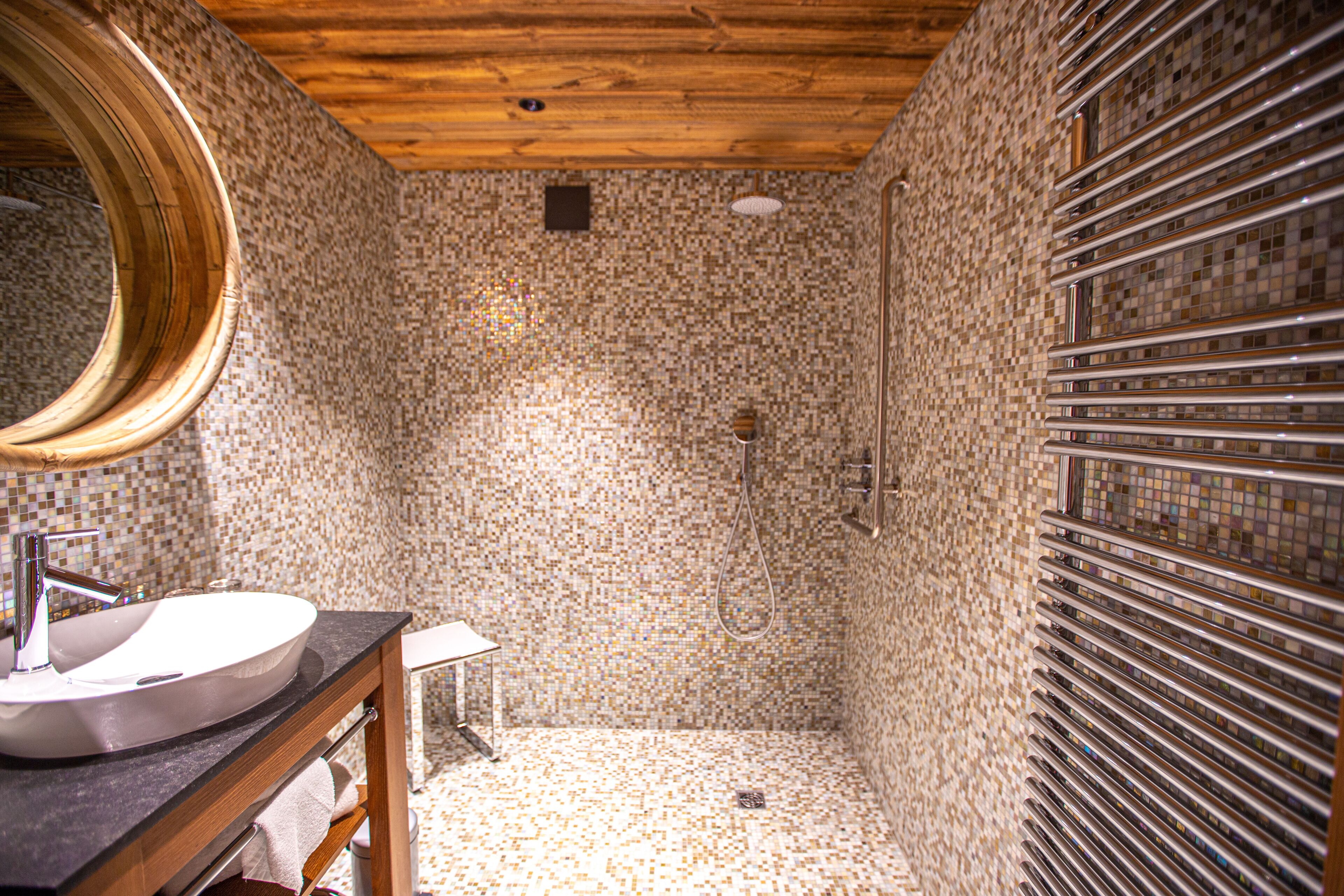 A bathroom with a mosaic tile shower, a wooden vanity with a round mirror, and a tiled floor.