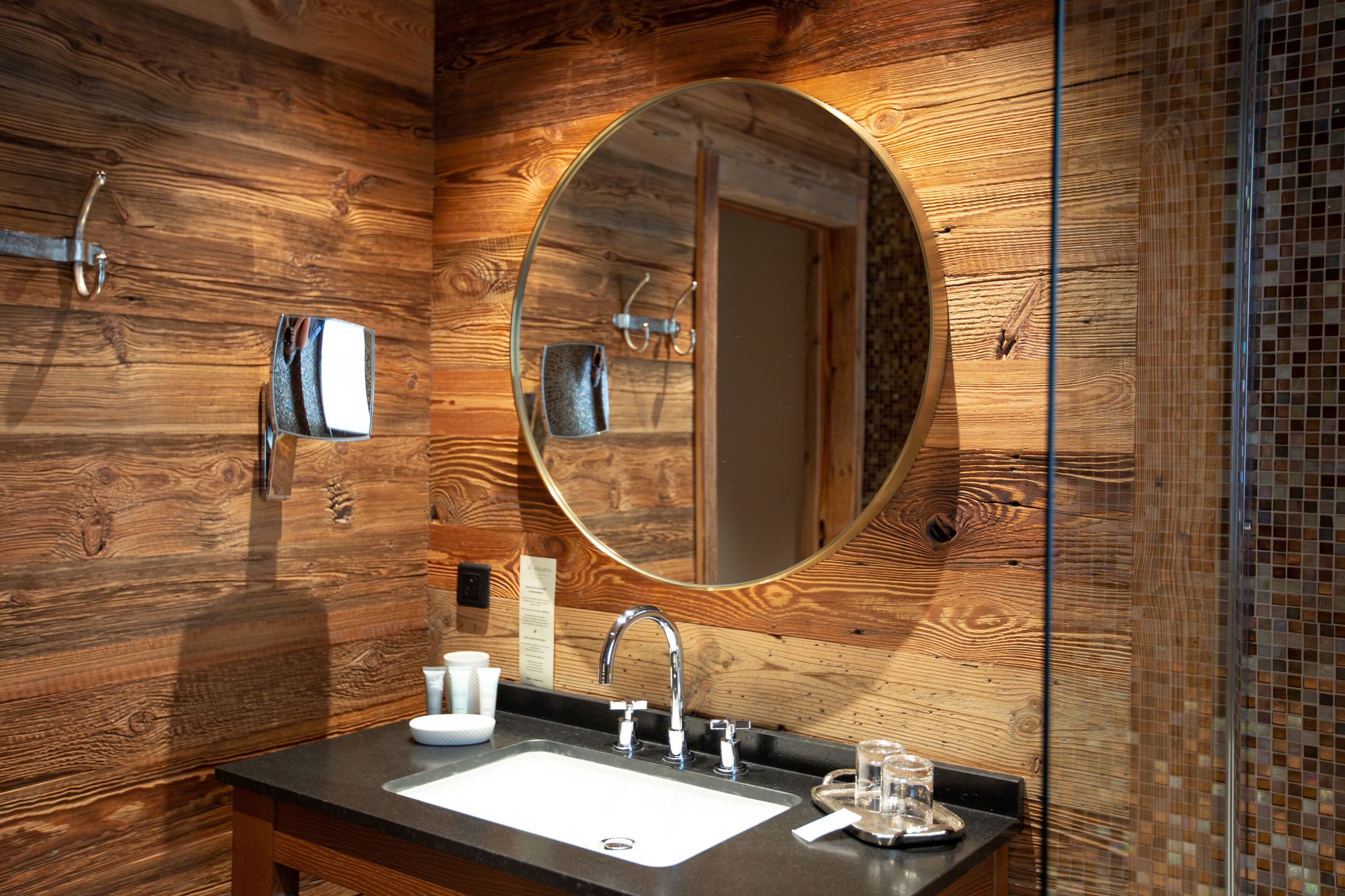 Een badkamer met een houten wand, een ronde spiegel, een wastafel en een glas water op het aanrecht.