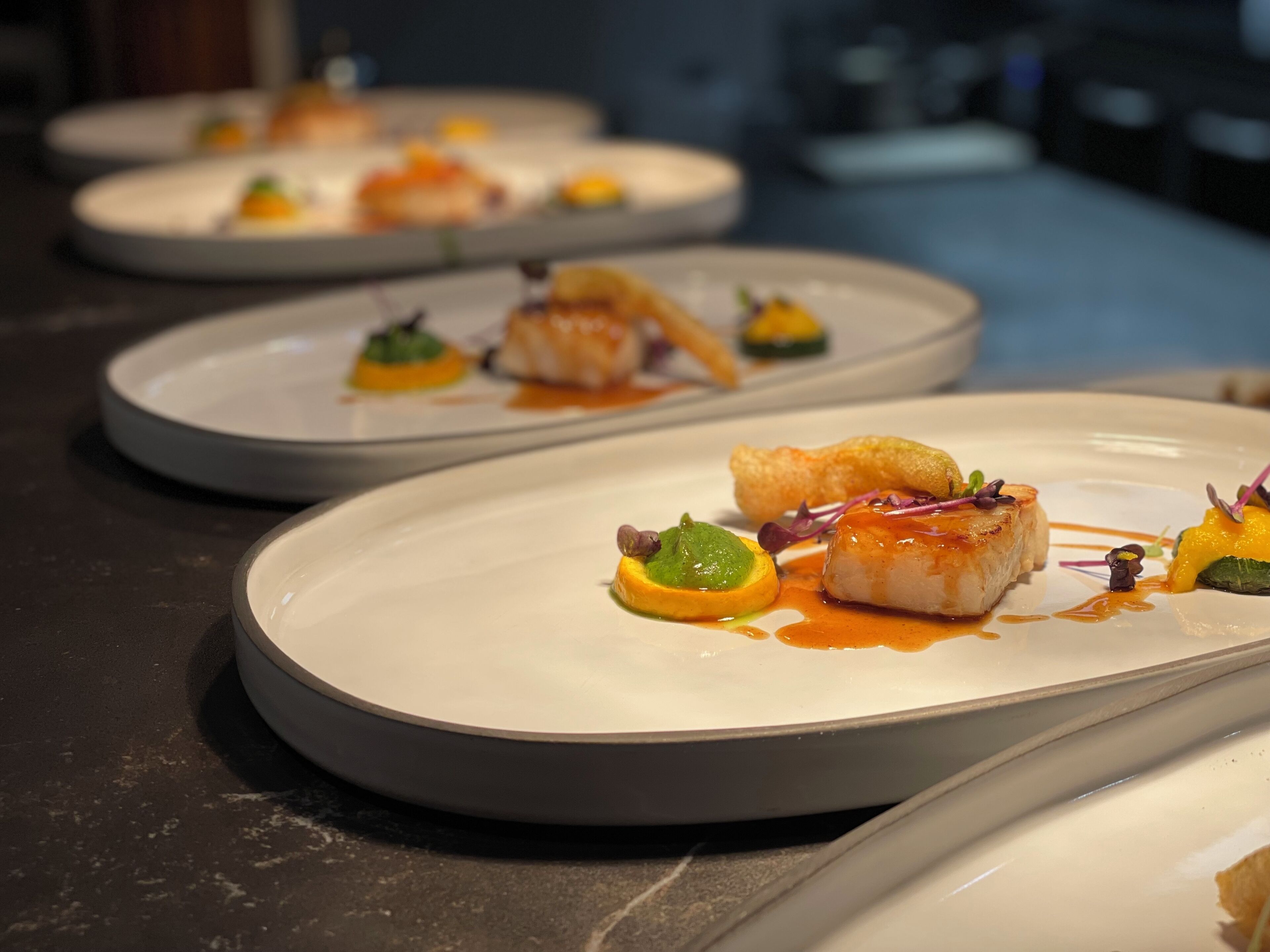 A series of elegantly plated dishes with a focus on seafood and vegetables, arranged on a dark marble countertop.