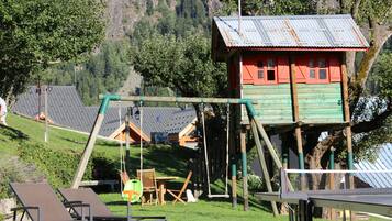 A wooden playhouse with a swing set, a small table, and a chair in a grassy area with a mountain backdrop.