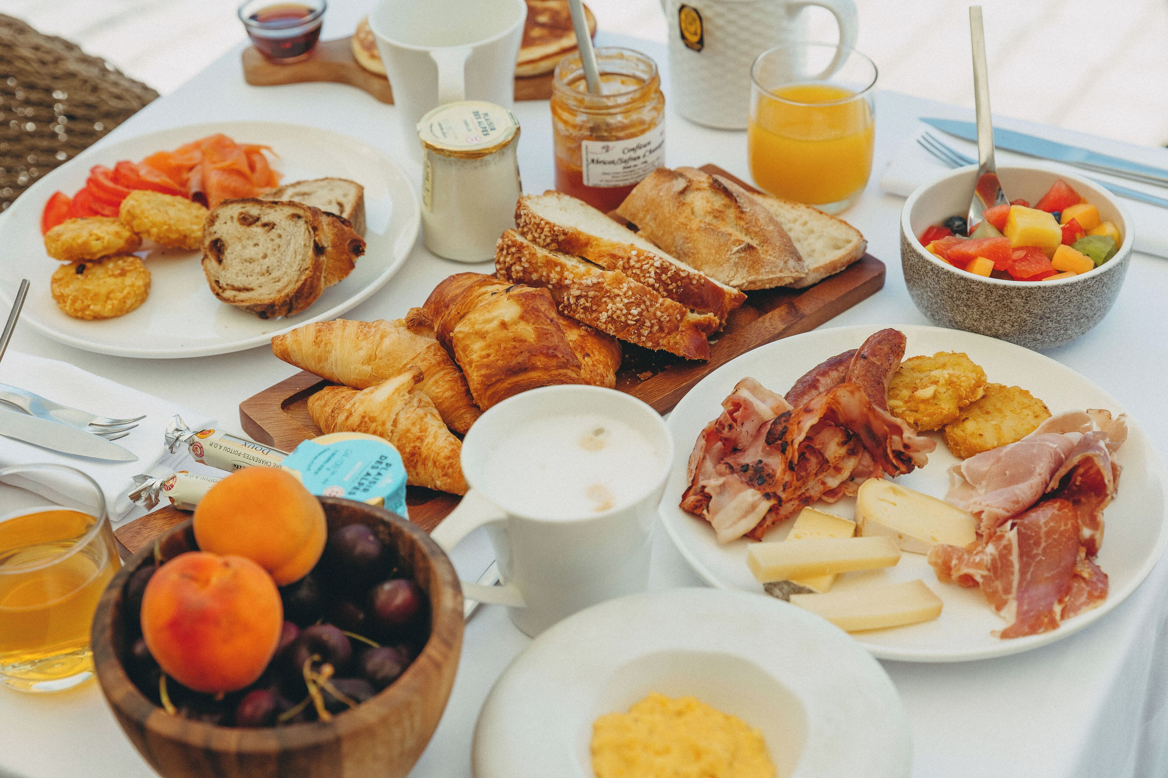 Una mesa de desayuno con diversos alimentos, como pan, bollería, queso, jamón y frutas.