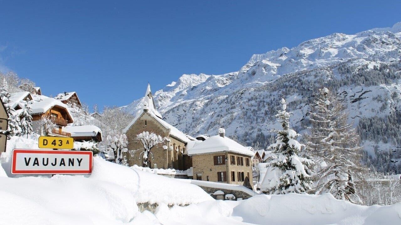 Un pueblo cubierto de nieve en la montaña con edificios de estilo chalete tradicional, cielo despejado y un cartel que indica Vaujany.