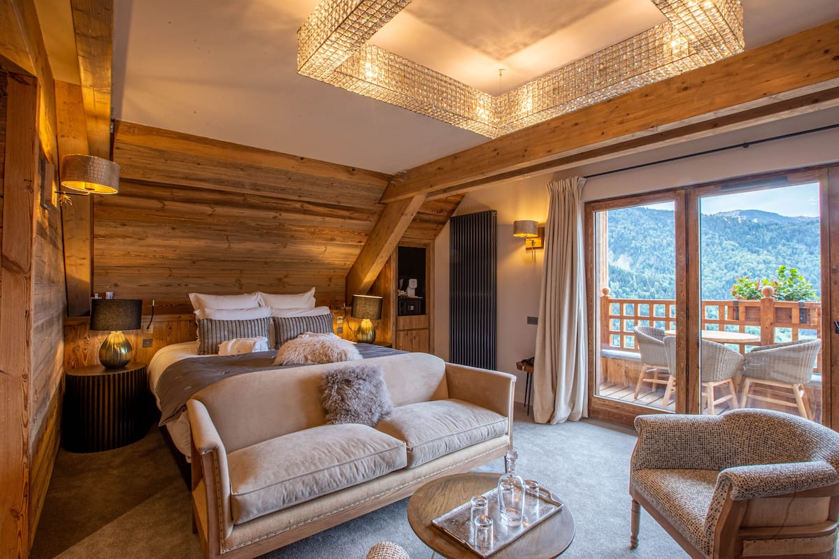A cozy log cabin bedroom with a large bed, a sofa, and a balcony with a wooden deck and chairs.