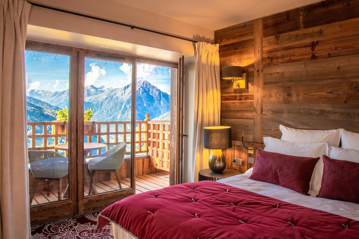 A bedroom with a large bed, a wooden headboard, a nightstand with a lamp, and a view of mountains through a sliding glass door.