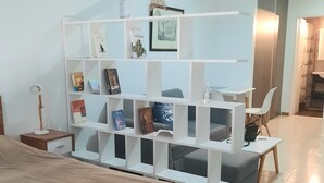Una camera da letto moderna con un letto, una libreria bianca piena di libri e oggetti decorativi, una scrivania con una sedia, e un bagno visibile sullo sfondo.