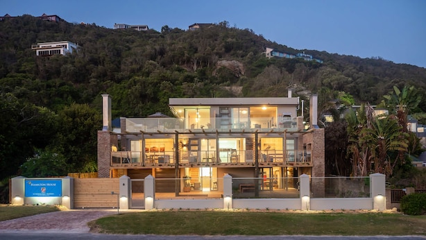 A modern two-story house with a glass balcony, surrounded by lush greenery and hills. - Hamilton House (Wilderness)