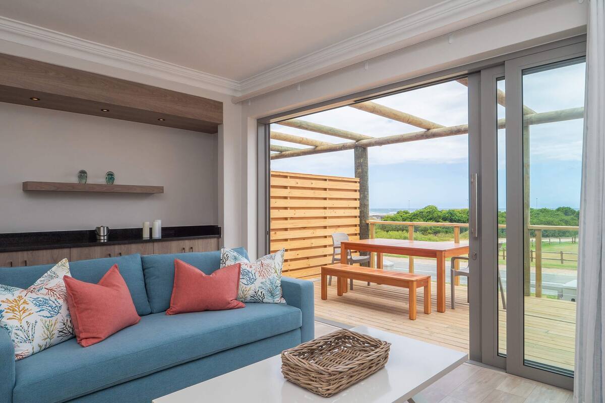 A modern living room with a blue sofa, a wooden table, and a view of the outdoors through large sliding doors.