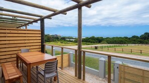 Une terrasse en bois avec une table et des chaises, donnant sur une route et une zone herbeuse, d’où l’on peut admirer l’océan.