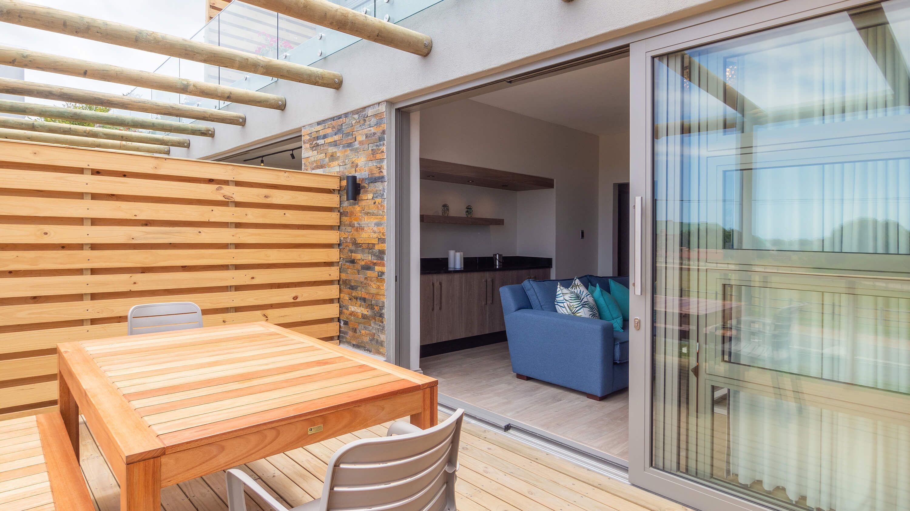 A wooden deck with a table and chairs, a blue sofa, and a kitchen area visible through an open door.