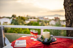 Et bord med rødt dug, to glas rødvin, et stearinlys og en blomsteropsætning på en balkon med udsigt over et smukt landskab.