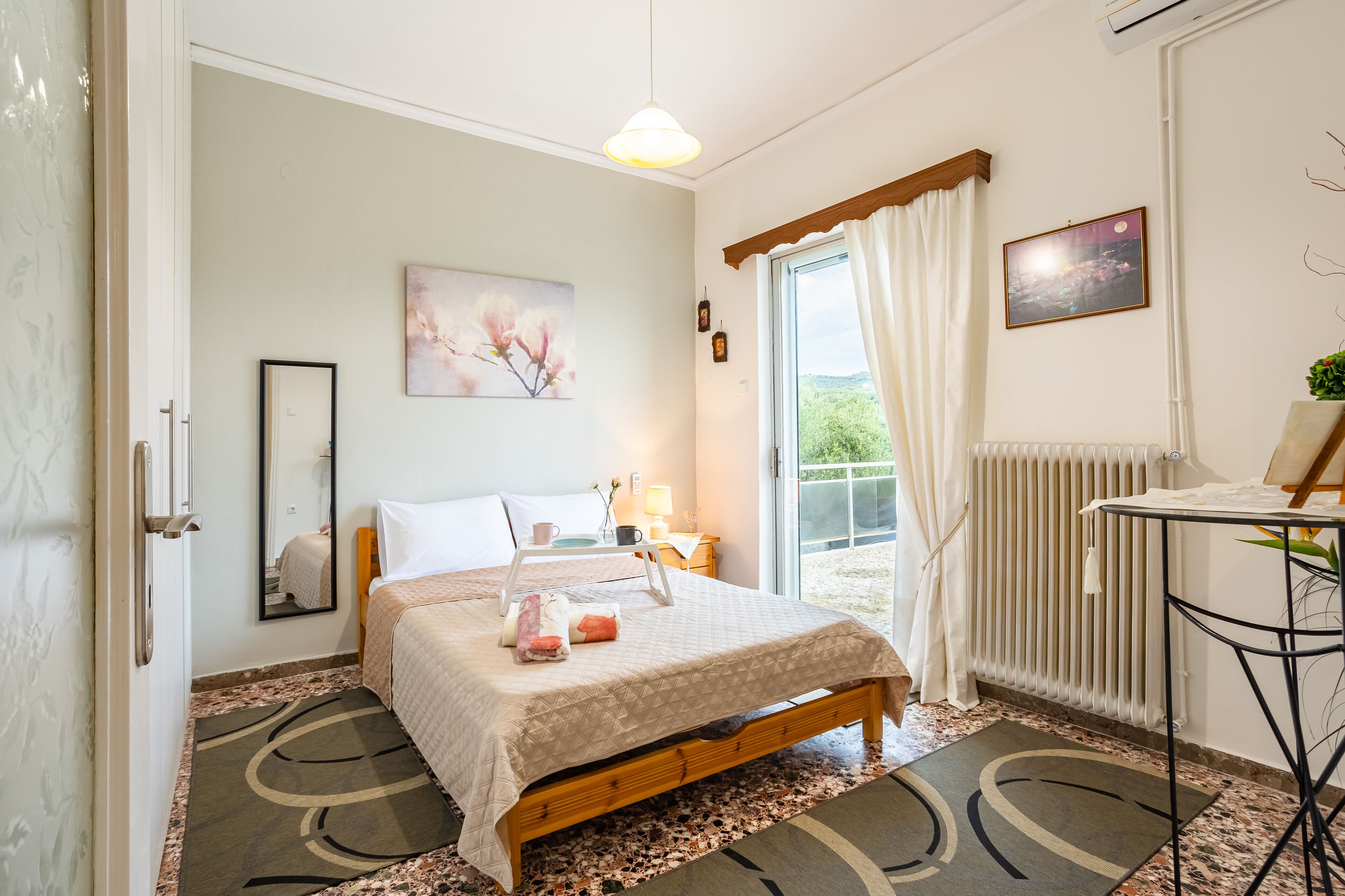 A bedroom with a bed, a bedside table, a window with curtains, a radiator, and a painting on the wall.