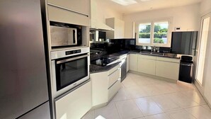 A modern kitchen with stainless steel appliances, white cabinetry, and a large window providing natural light. - Mediterranean-style villa with pool (Agde)