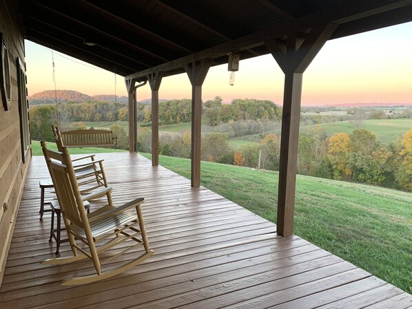 A wooden deck with rocking chairs overlooking a rural landscape during sunset.