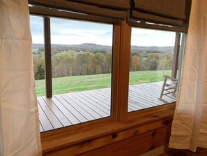 A wooden deck with a rocking chair, large windows with curtains, and a view of a green field and trees.