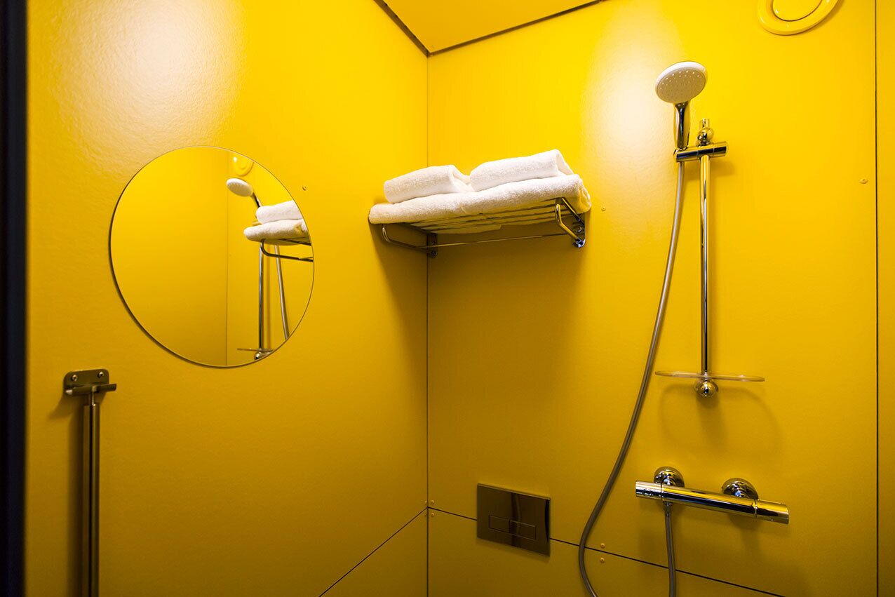 Une salle de bain avec un mur jaune, un miroir rond, un porte-serviettes avec des serviettes pliées et une douche équipée d’un pommeau de douche à main.