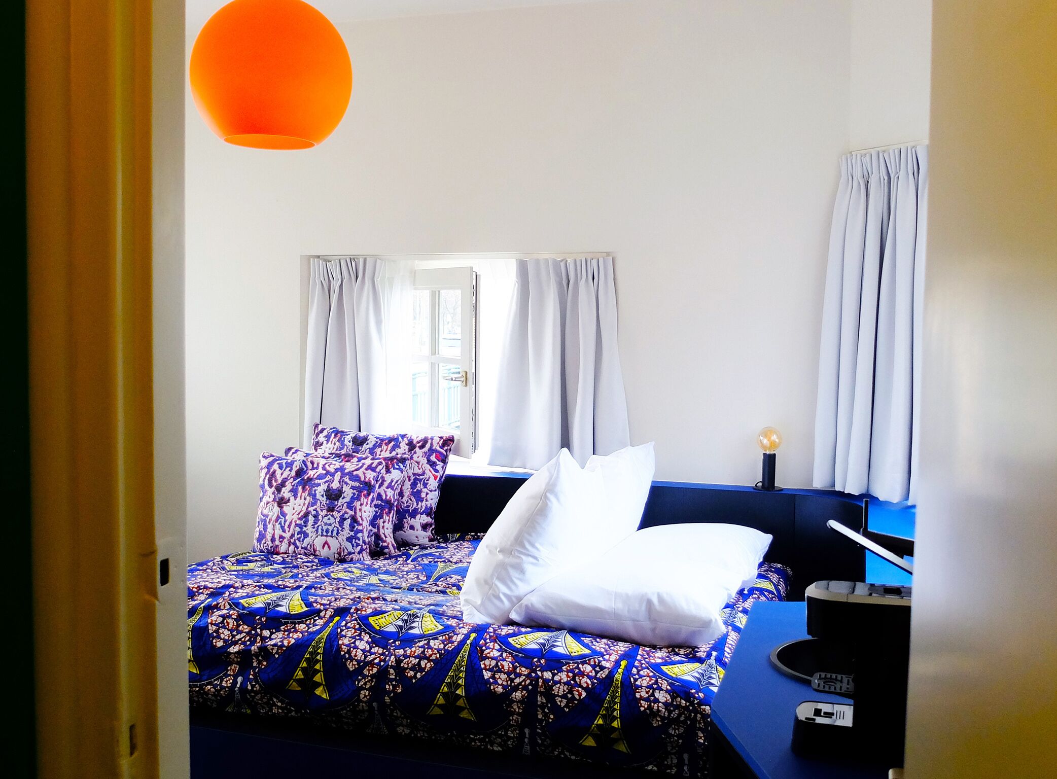 A bedroom with a bed, white pillows, a blue headboard, a nightstand with a lamp, and a window with curtains.