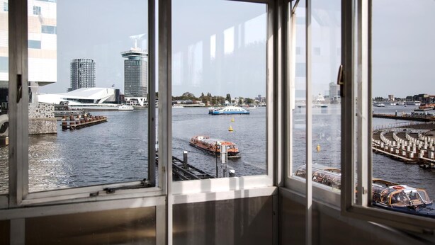Blick durch große Fenster auf einen Fluss mit Booten, ein modernes Gebäude und eine Brücke.