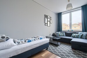 A modern living room with a sofa, a bed, and a coffee table. There are framed pictures on the wall and a window with curtains. - Labo Apartment Tamka (Warsaw)