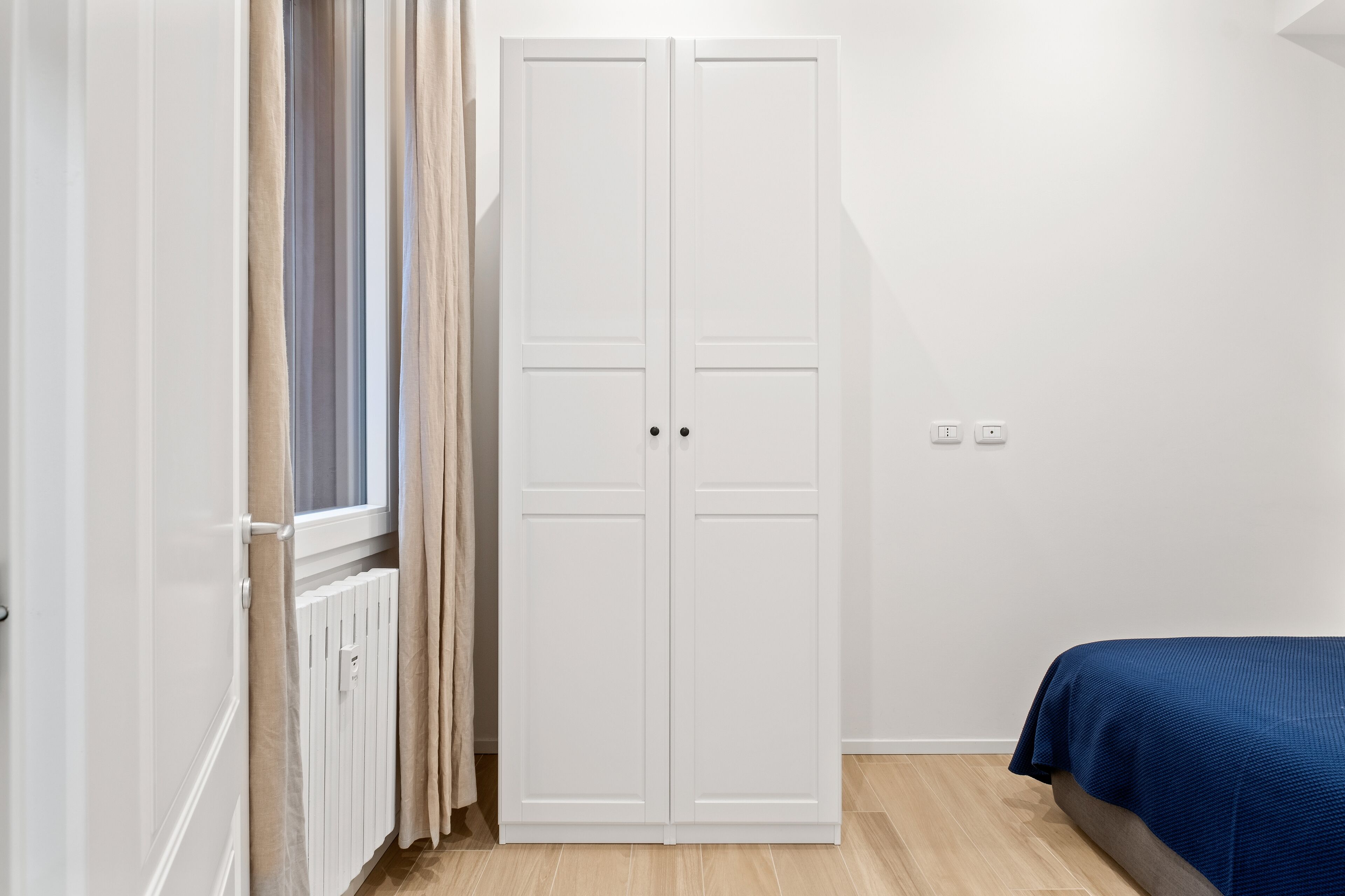 Een slaapkamer met een witte kast, een raam met gordijnen, een radiator en een bed met een blauwe spreidek.