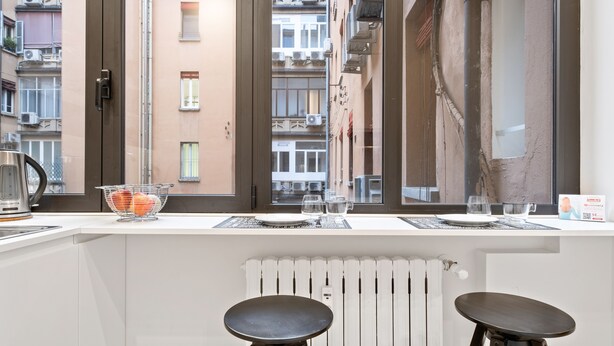 A modern kitchen with a white countertop, two stools, and a window overlooking a cityscape. - Piazza Maggiore White Apartment (Bologna)