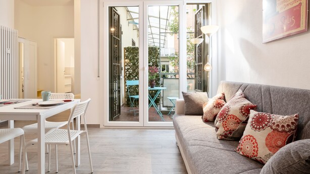A modern living room with a sofa, a dining table with chairs, and a sliding glass door leading to an outdoor area with a table and chairs. - Giardini Margherita Monolocale con Balcone (Bologna)
