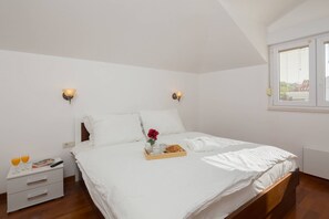 Un lit bien fait, avec du linge de lit blanc, une table d’appoint avec un livre, une télécommande et une fleur. Il y a deux lampes fixées au mur et une fenêtre avec des stores.