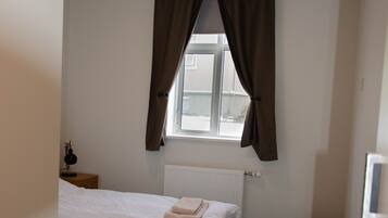 Una habitación pequeña y limpia con una cama, una mesita de noche, una ventana con cortinas y un radiador.