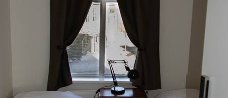 A small, single-room hotel room with two beds, a desk, a lamp, and a window with curtains.