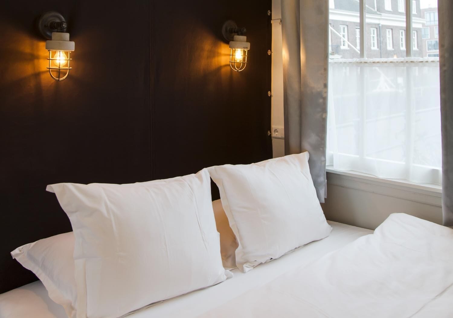 A hotel room with a dark wooden headboard, two white pillows, and two wall-mounted light fixtures.