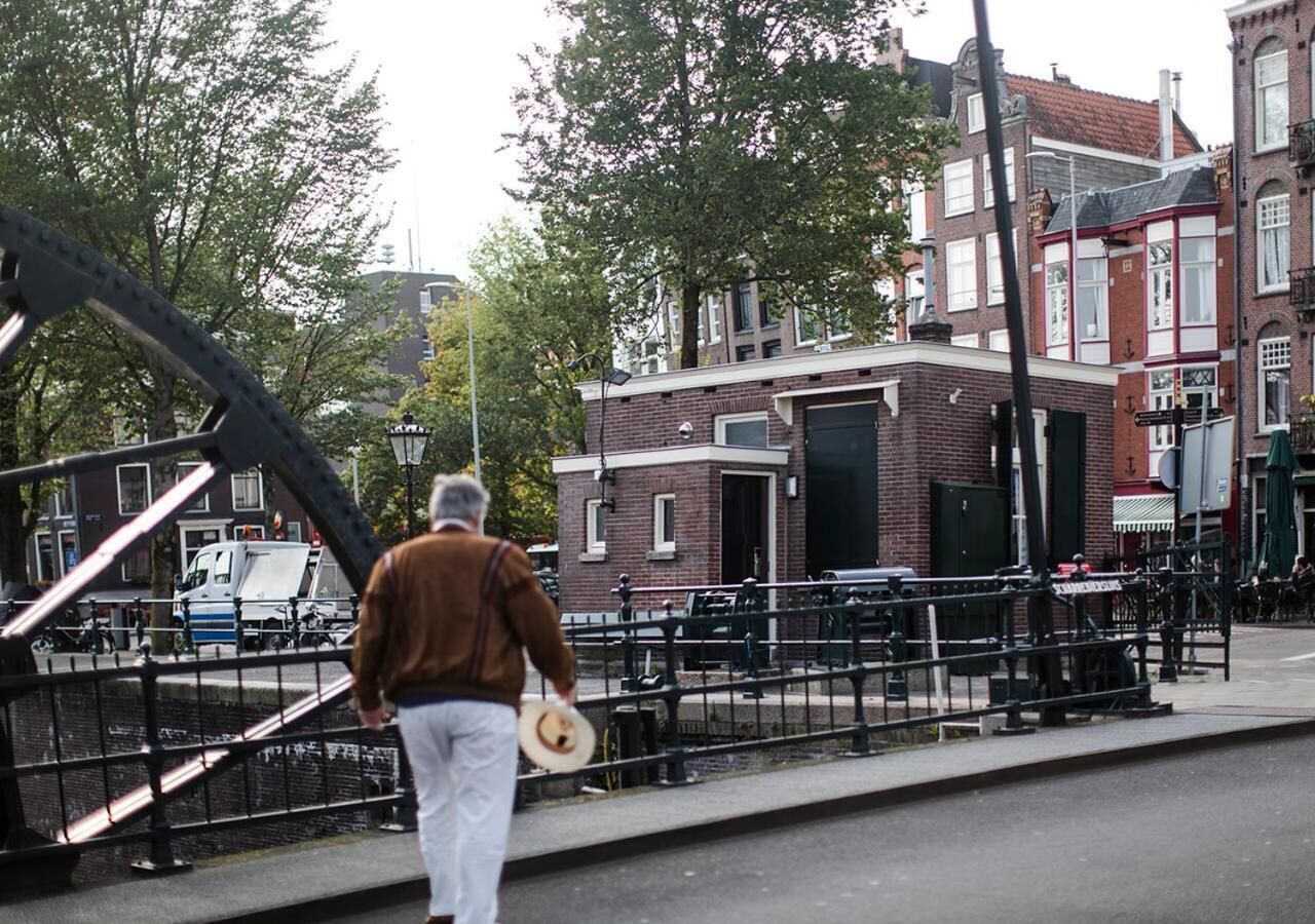 Un hombre caminando sobre un puente, con sombrero, frente a un edificio de ladrillo con una puerta negra, y una hilera de casas de ladrillo rojo al fondo.