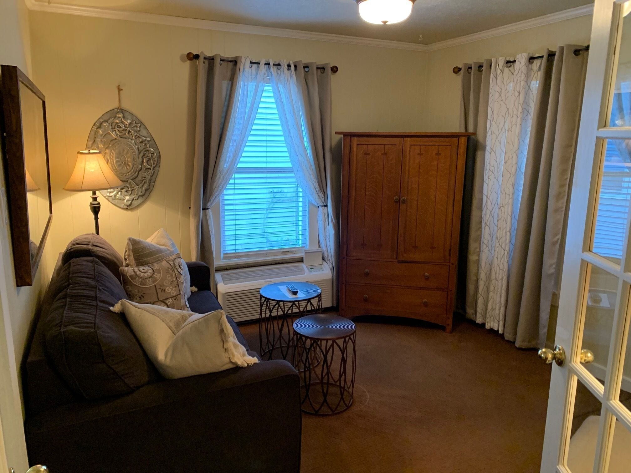 Una sala de estar acogedora con un sofá, una lámpara, una ventana con cortinas, un mueble de madera y una mesita.