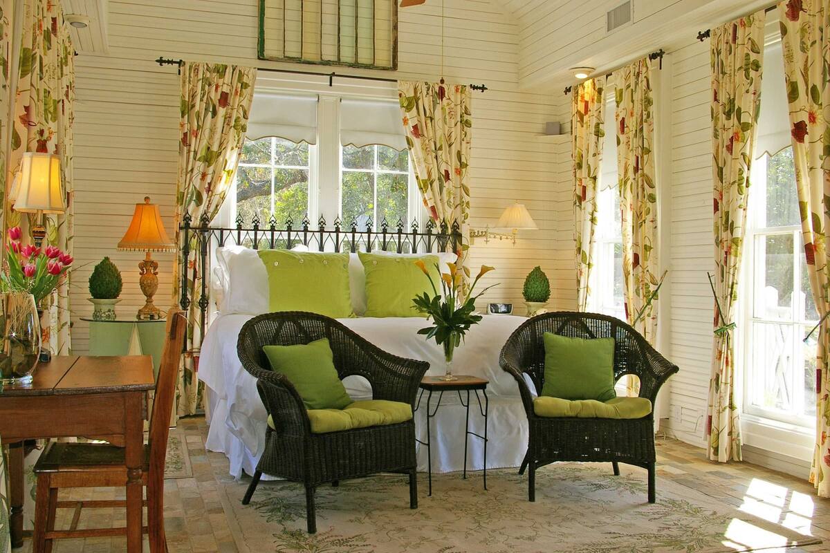 A cozy bedroom with a bed, two wicker chairs with green cushions, a small table, and a vase with flowers.