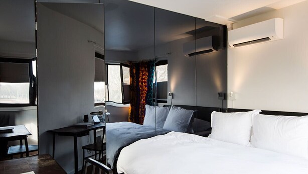 A modern hotel room with a large bed, a desk, and a chair. There is a window with curtains, an air conditioning unit, and a wall-mounted TV. - SWEETS - Meeuwenpleinbrug (Amsterdam)