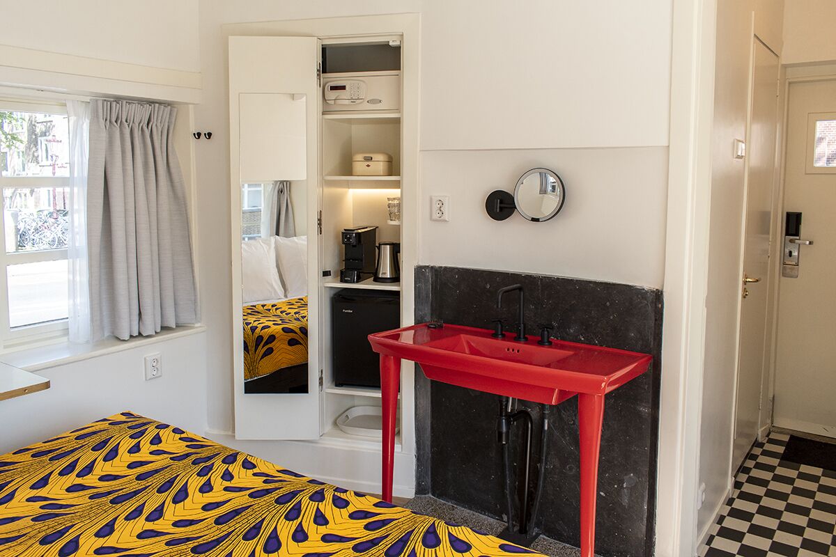 Une pièce de taille modeste comprenant un lit, un lavabo rouge, un petit coin cuisine et une fenêtre avec des rideaux.