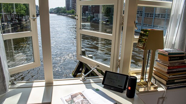 A desk with a laptop, a book, and a stack of books near a window overlooking a river. - SWEETS - Van Hallbrug (Amsterdam)