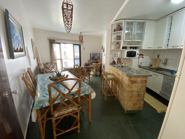 A dining area with a table set for four, surrounded by wooden chairs. A kitchen with white cabinets and a microwave is visible to the right. - Apt well decorated and complete 50 meters from the beach + beach service (Vila Clais)