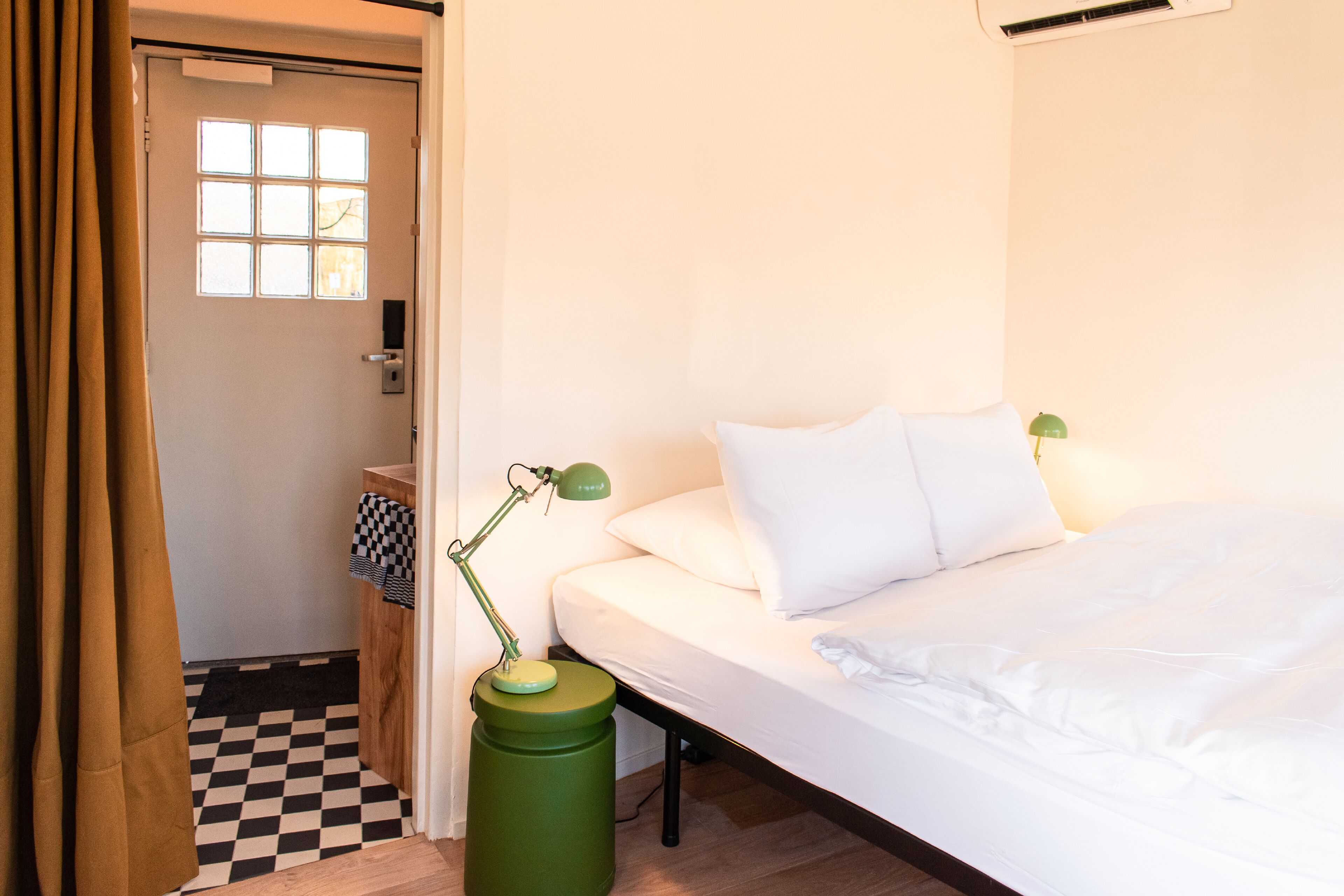 A hotel room with a bed, a desk with a lamp, a window, and a door.