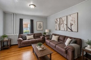 Un salón con dos sofás, una mesa de centro, un radiador, una ventana con cortinas y decoración en la pared.