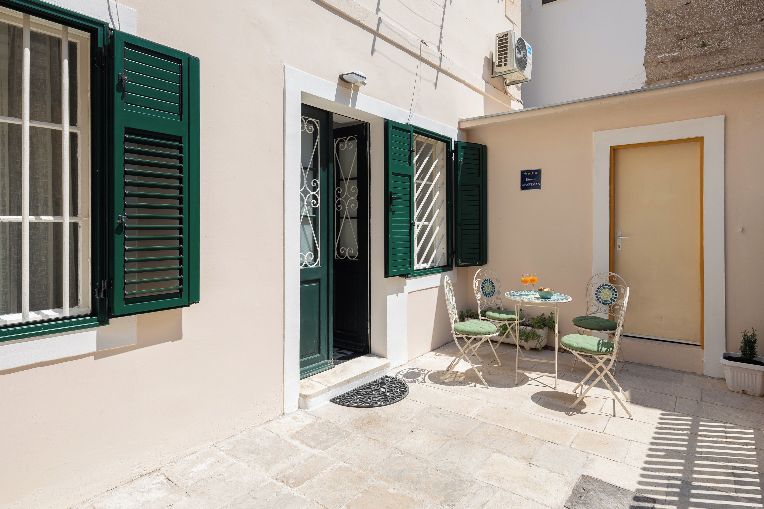 A small courtyard with a table and two chairs, a door, and a window with green shutters.