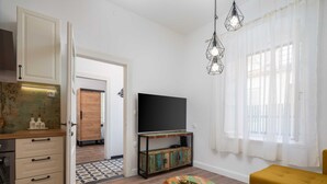 Ein Wohnzimmer mit Fernseher, einem gelben Sofa, einem Holztisch und einer Küchenzeile mit Schränken und Arbeitsplatte.