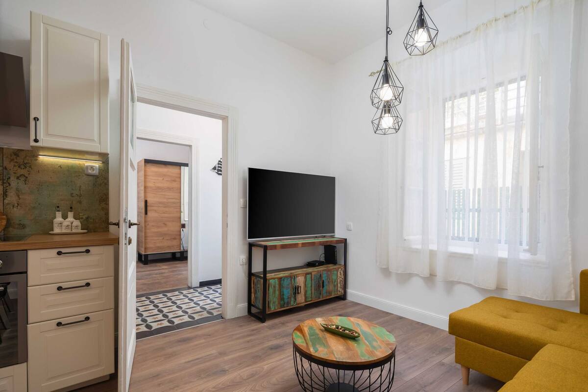 A living room with a TV, a yellow sofa, a wooden coffee table, and a kitchen area with cabinets and a countertop.