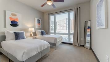 A hotel room with two beds, a ceiling fan, large windows, and artwork on the walls.